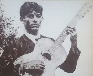Foto antiga, em preto e branco, com sinais de desgaste, de um homem jovem de cabelos escuros e lisos, segurando um violão inclinado para cima.