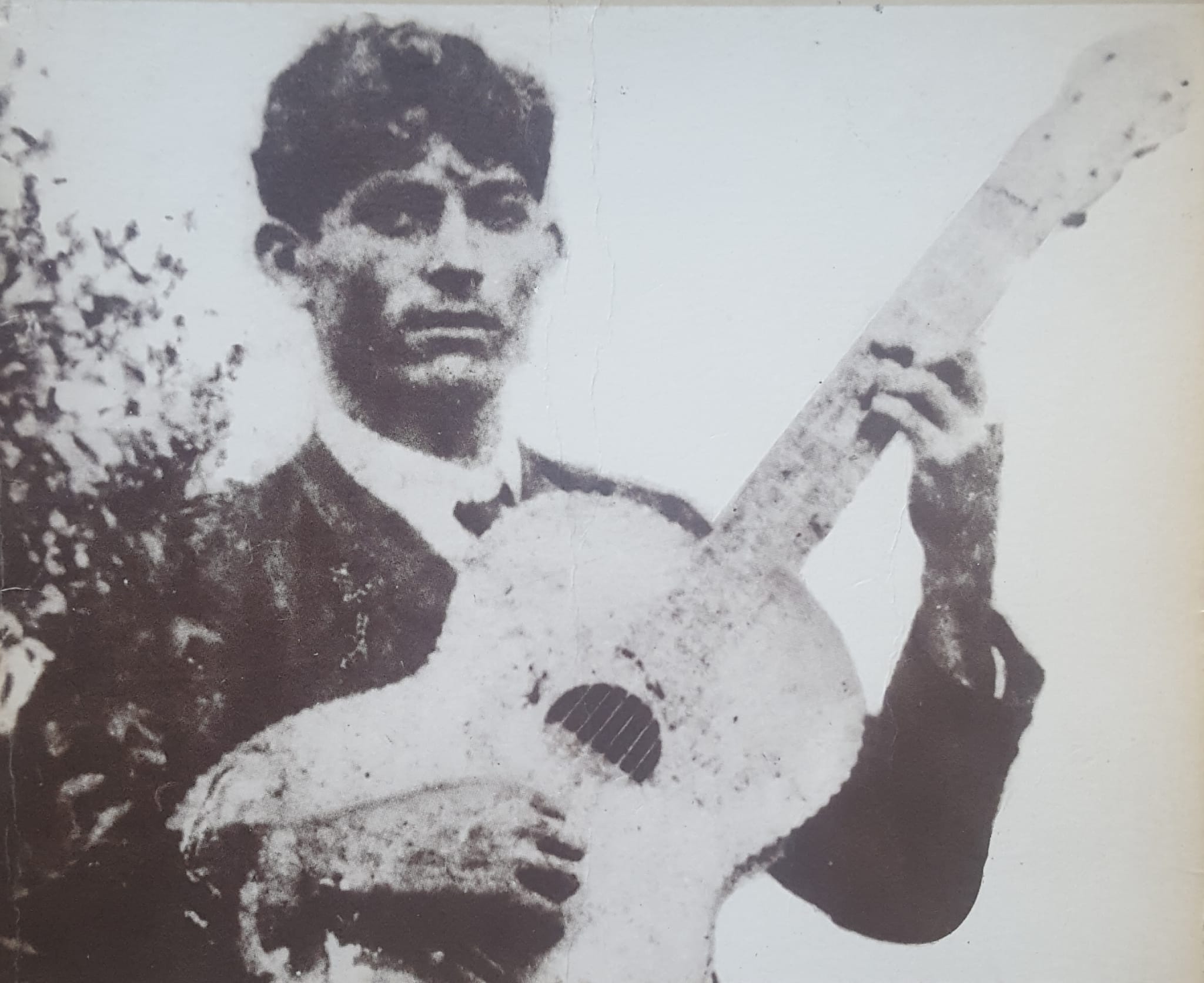 Foto antiga, em preto e branco, com sinais de desgaste, de um homem jovem de cabelos escuros e lisos, segurando um violão inclinado para cima.