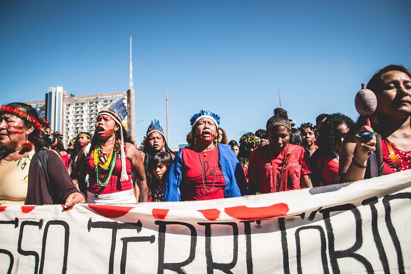 Grupo de mulheres indígenas em marcha sobre Brasília, sob céu azul sem nuvens, por trás de uma faixa branca com letras pintadas de preta onde se lê parte da palavra "território".