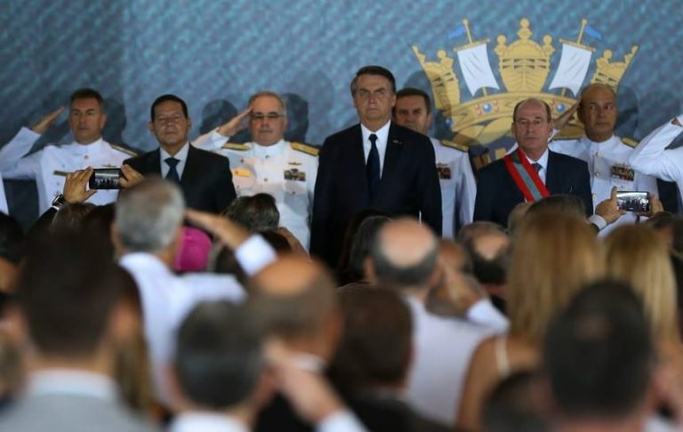 Em palaque, Jair Bolsonaro está perfilado, de paletó e gravata escuros, recebendo a continência do Alto Comando da Marinha, composto por homens fardados de uniformes brancos com medalhas. Em primeiro plano, desfocadas, dezenas de pessoas de costas olham para o palco também batendo continência.