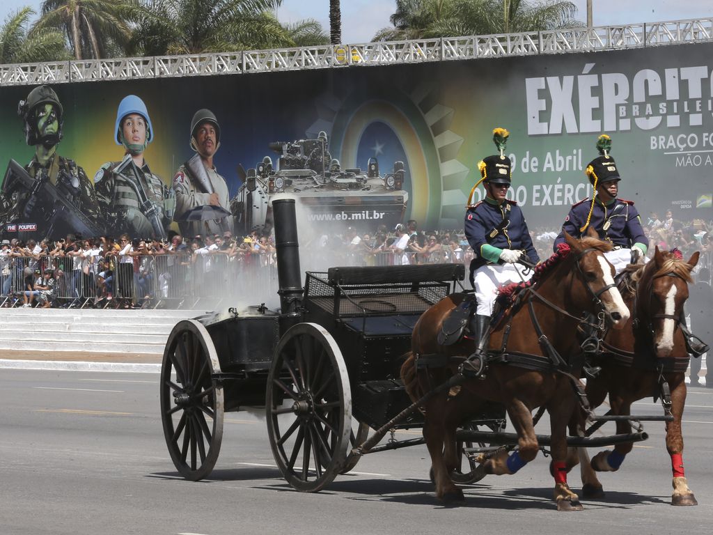 À frente de um enorme painel em homenagem ao Exército, dois cavaleiros fardados com uniformes de gala, desfilam com seus cavalos puxando uma carroça que carrega antigo artefato militar que solta fumaça