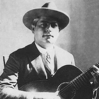 Foto em preto e branco de homem branco, com chapéu, semblante sério, segurando violão.