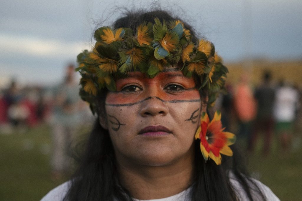 Close de mulher indígena com pintura na cor alaranjada em torno dos olhos, adorno tradicional verde e amarelo na cabeça e flor vermelha com bordas amarelas usada como brinco na orelha esquerda.