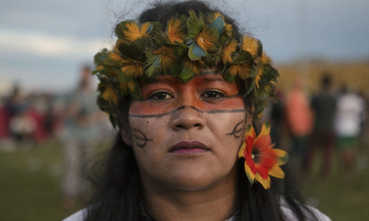 Close de mulher indígena com pintura na cor alaranjada em torno dos olhos, adorno tradicional verde e amarelo na cabeça e flor vermelha com bordas amarelas usada como brinco na orelha esquerda.