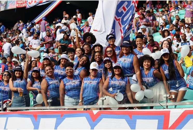 Grupo de mulheres vestindo azul, vermelho e branco em meio a uma multidão em estádio de futebol.