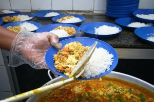 Mão segura um prato azul onde está sendo colocada uma porção de arroz e outro alimentos. Ao fundo, outros pratos da mesma cor, todos com alimento, em cima de uma balcão.