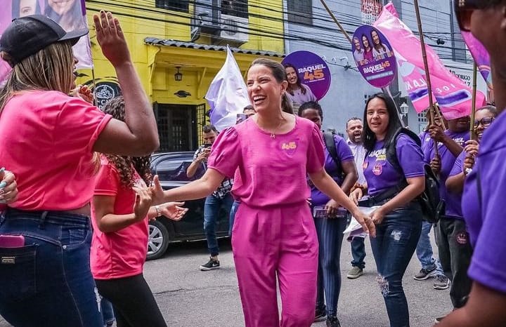Raquel Lyra: mulher branca de cabelos presos, usando blusa e calça lilás, com sorriso aberto, cumprimenta outras mulheres que carregam cartazes de sua campanha eleitoral no meio de uma rua.
