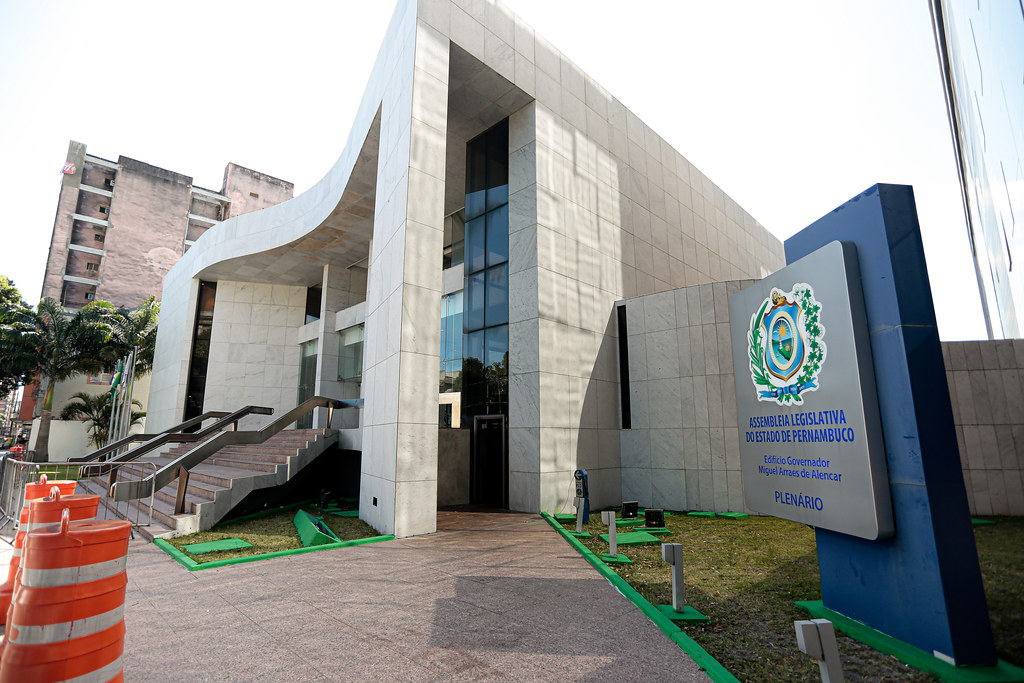 Foto diurna da fachada do prédio sede da Assembleia Legislativa de Pernambuco, em granito, cinza, com brasão do Estado de Pernambuco e nome do local no lado direito da imagem