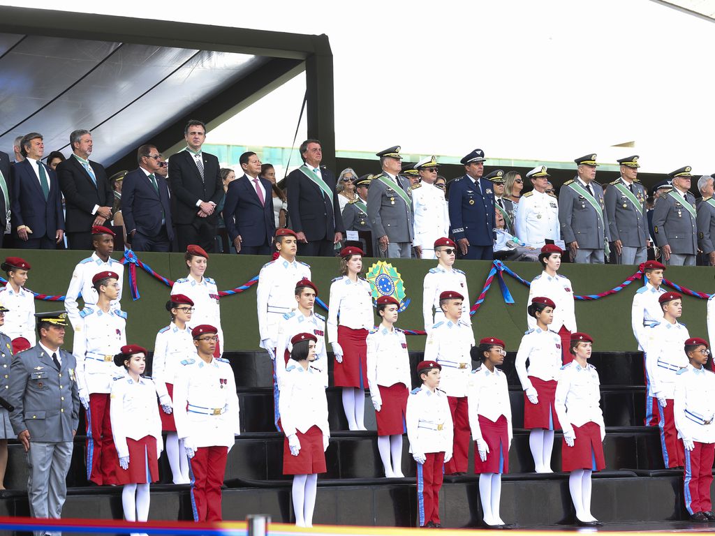 No alto de um palaque, dezenas de homens fardados perfilados, tendo ao centro Jair Bolsonaro. Abaixo do palanque, estão jovens, rapazes e moças, usando uniformes de gala com calças vermelhas e blusões brancos.