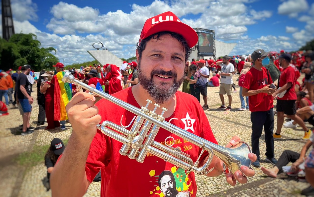 Foto de homem branco, usando camisa vermelha e boné vermelho, ambos de Lula. Ele segura um trompete. Ao fundo muitas pessoas.