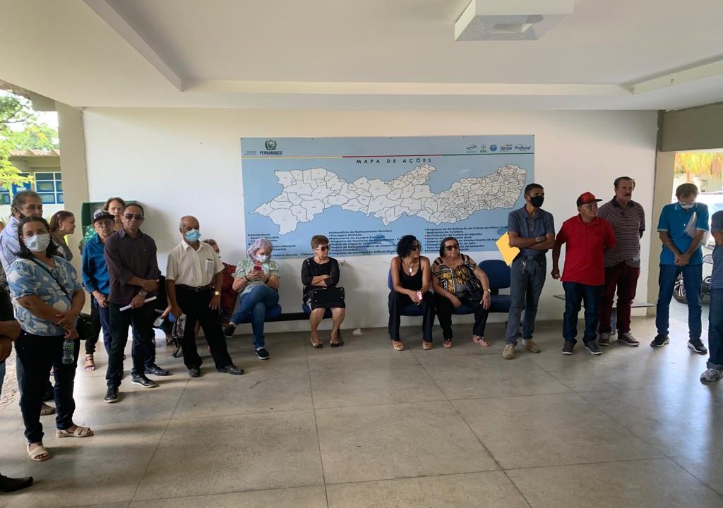 Grupo de homens e mulheres de meia idade reunidos em semicírculo em salão com iluminação natural. Por trás deles, mapa de Pernambuco.