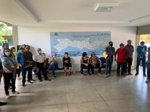 Grupo de homens e mulheres de meia idade reunidos em semicírculo em salão com iluminação natural. Por trás deles, mapa de Pernambuco.