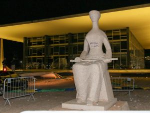 Foto noturna da fachada do STF, sem iluminação, onde se vê em primeiro plano escultura simbolizando a Justiça com pichações feitas por bolsonaristas