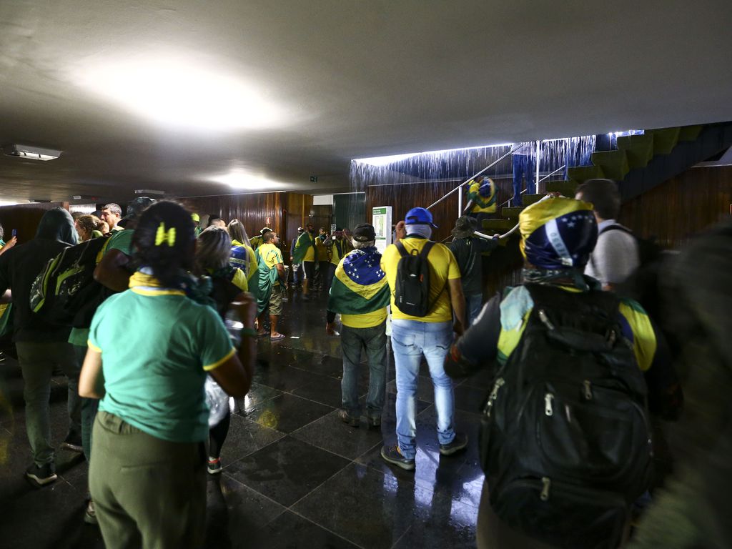 grupo de pessoas vestindo camisas amarelas e usando bandeiras do Brasil depredam moveis de salão com piso de granito e paredes forradas de madeira.