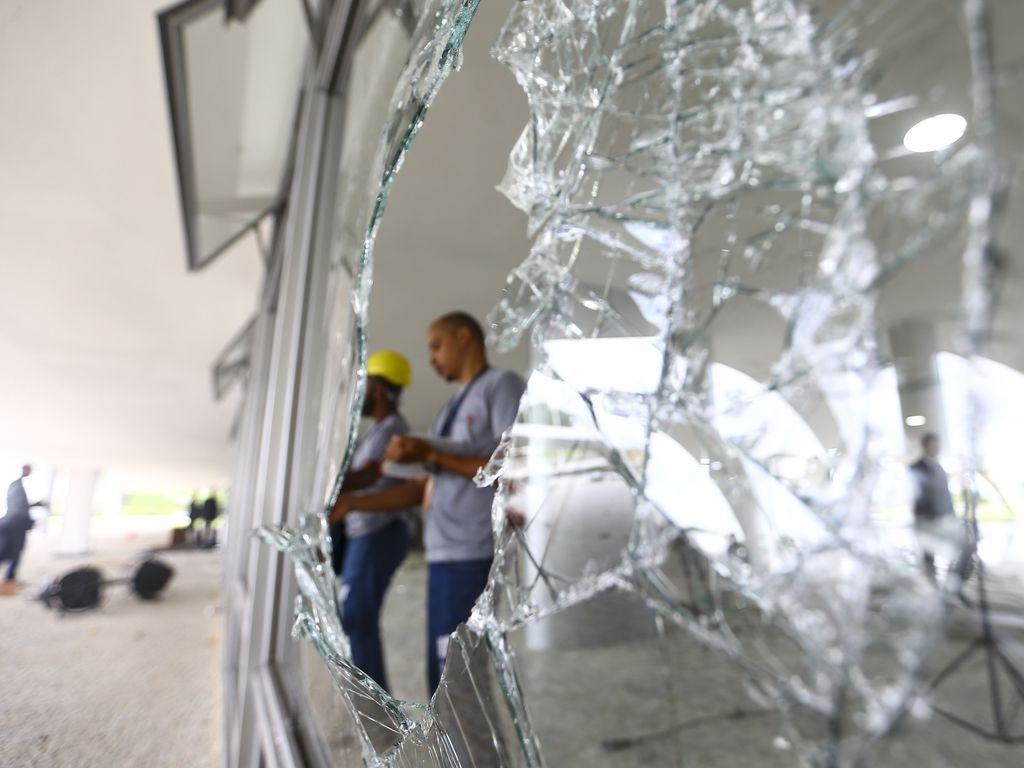 Do buraco de uma janela estilhaçada, se vê dois operários fazendo reparos no interior do Palácio do Planalto