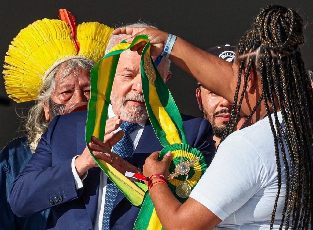 Mulher negra, de tranças e camiseta branca, coloca a faixa presidencial verde e amarela no corpo de Lula, sob o olhar do cacique Raoni, que usa grande cocar amarelo com pena vermelha ao fundo da foto.