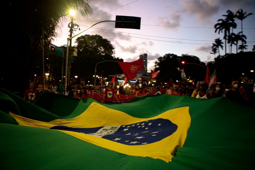 Sob a luz do por do sol, multidão usando em sua maioria camisas vermelhas leva uma grande bandeira do Brasil.