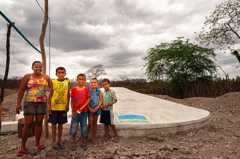 Mulher acompanha de quatro crianças pequenas em "escadinha", do mais alto ao mais baixo, da esquerda para a direita, posam em frente à cisterna (grande tanque de cimento branco) construída sobre solo seco, com árvores ao fundo, à direita da imagem.