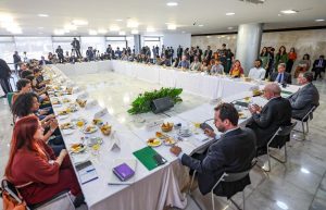 grande mesa retangular, coberta com pano branco, com dezenas de pessoas, tendo Lula ao centro, no lado direito da imagem.