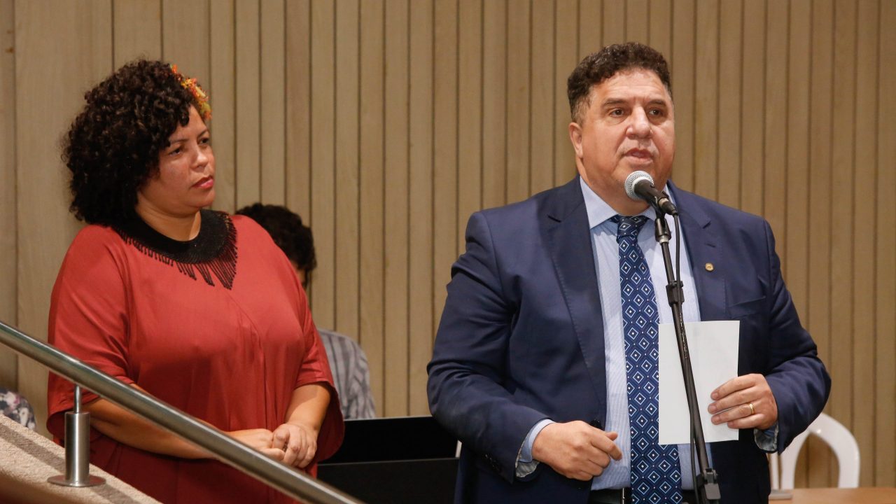 No plenário da Assembleia Legislativa, o pastor Cleiton Collins (homem pardo, de cabelos curtos escuros, paletó cinza azulado e gravata azul) discursa, sendo observado por Dani Portela (mulher negra de vestido cor de tijolo com flor amarela nos cabelos crespos escuros), que está posicionada por trás dele, em posição de fila de espera.