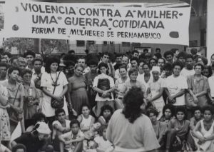 Foto em preto e branco com grande grupo de mulheres à frente de faixa branca pintada com a frase "Violência contra a mulher, uma guerra cotidiana".