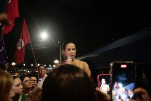 À noite e ao ar livre, Raquel Lyra discursa usando microfone em meio à multidão, com pessoas que fazem foto dela usando celulares, tendo uma bandeira vermelha do MST ao fundo.