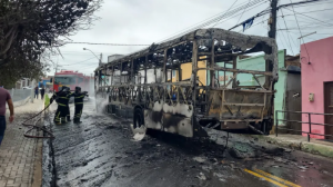 No meio de uma rua estreita, durante o dia com céu nublado, carroceria de ônibus completamente destruído pelo fogo, ainda esfumaçando, com bombeiros apontando mangueira d'água em direção a uma das extremidades do veículo