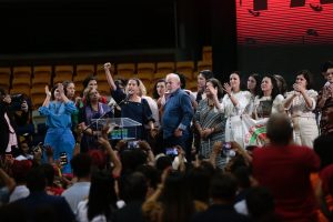 Cercados de ministros no palco, Lula, de camisa azul, está ao lado de Raquel Lyra, de vestido escuro com o braço direito levantado