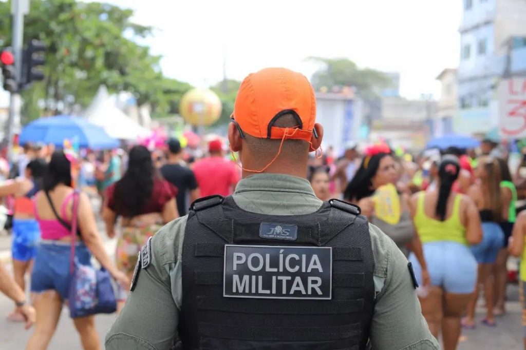 Em primeiro plano, soldado da PM de costas, usando boné laranja e colete preto com a inscrição Polícia Militar. Ao fundo, bastante desfocadas, mulheres usando shorts e camisetas coloridas na rua ao livre.