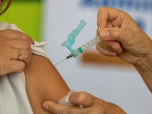 Imagem de duas mãos aplicando vacina em braço de uma pessoa não identificada, cuja manga da camisa de cor branca está levantada.