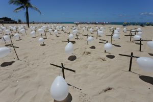 dezenas de cruzes pretas com balões de ar branco fincadas na areia da praia, com coqueiro à esquerda da imagem e o mar ao fundo, sob o azul do céu.