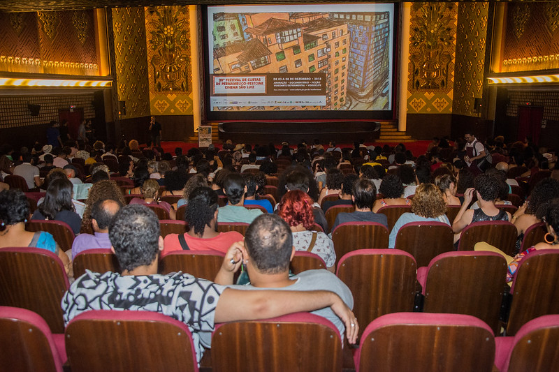 Sala de cinema, com foto focando a tela colorida, com pessoas na plateia de costas e sentadas em poltronas forradas na cor vermelha e encostos de madeira.