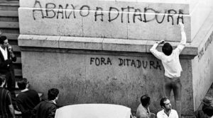 Foto preto e branco de rapaz de camisa branca e calça comprida grafitando as palavras abaixo a ditadura em uma larga coluna de um prédio, sendo observado por outras pessoas. Todos estão de costas para a câmera.