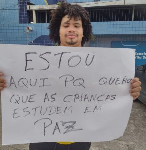 Homem negro, jovem de fartos cabelos encaracolados e bigode ralo, segura cartaz com a frase "Estou aqui pq quero que as crianças estudem em paz"