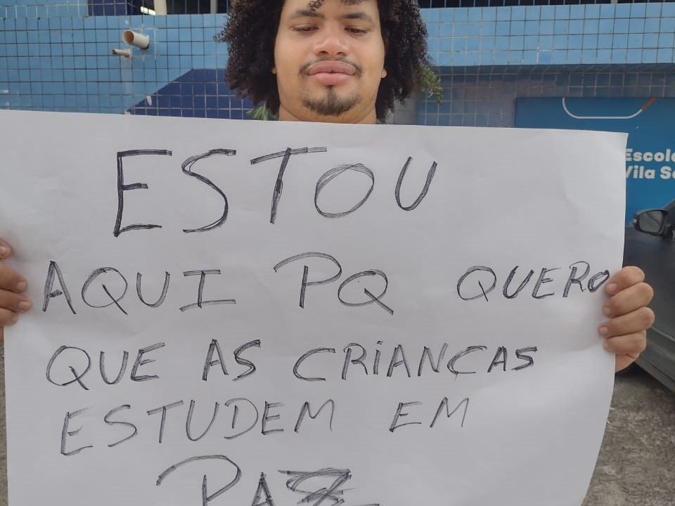 Homem negro, jovem de fartos cabelos encaracolados e bigode ralo, segura cartaz com a frase "Estou aqui pq quero que as crianças estudem em paz"
