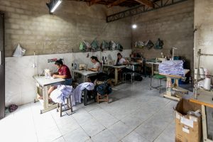 Em uma oficina de trabalho de parede de tijolos sem reboco e piso de cerâmica dr cor clara, três mulheres de cabelos pretos estão debruçadas sobre máquinas de costura instaladas em mesas enfileiradas.
