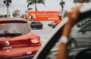 Em uma avenida, com sinal fechado, pessoas carregam faixa laranja com frases alusivas ao Dia Nacional de Enfrentamento ao Abuso e à Exploração Sexual das Crianças e Adolescentes: "Abuso sexual infantil é crime - Disque 100 e denuncie"