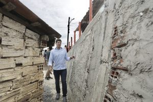 João Campos: homem branco jovem, de cabelos loiros, usa camisa branca de mangas compridas e calça jeans escura em uma viela entre um muro de arrimo e uma casa de tijolos aparentes.