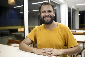 Henrique Vieira: homem negro, jovem, vestindo camisa amarela, sentado à mesa, sorrindo e olhando diretamente para a câmera.