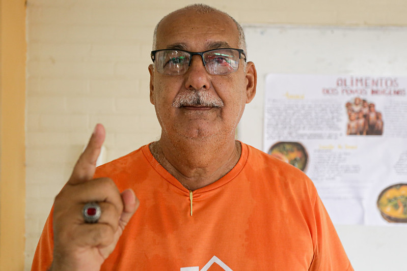Foto acima do peito de Wilson Lapa: homem branco, idoso, de óculos, bigode branco e careca, vestindo camisa laranja e com o dedo indicador da mão direita levantado.