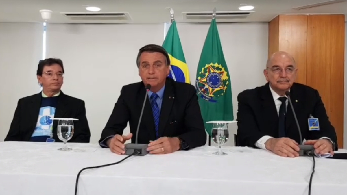 Médico Antônio Jordão, Jair Bolsonaro e o deputado Osmar Terra em live, sentados em uma mesa branca, com a bandeira do Brasil ao fundo.