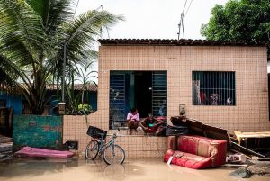 Casa com fachada de cerâmica rosa, com um sofá vermelho estragado pela água e outros entulhos, jogados junto ao portão da frente, em uma rua cheia de lama.