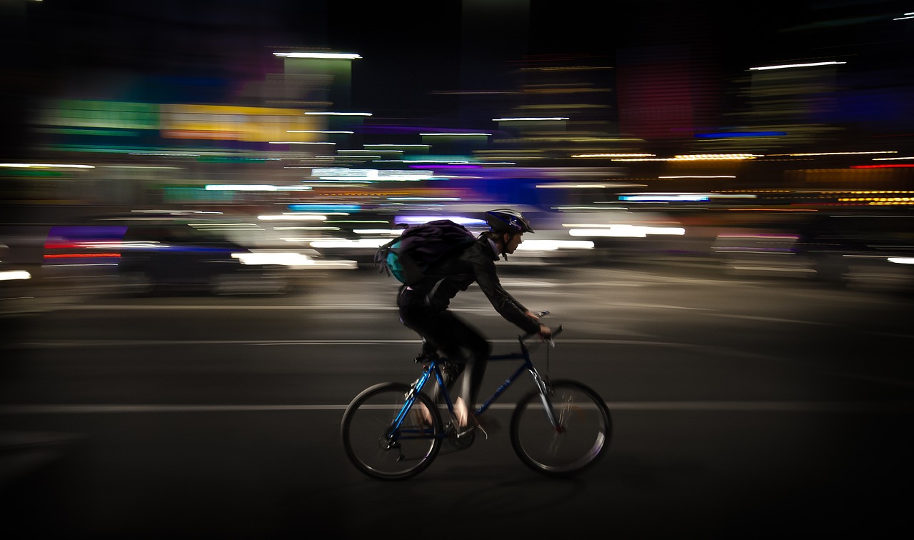 Foto noturna de ciclista com bolsa de entregador nas costas, em velocidade em uma avenida em que os carros e as luzes estão em segundo plano e fora de foco.