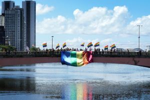 Em foto feita no leito do rio, vista da ponte Duarte Coelho, no centro do Recife, com pessoas erguendo pequenas bandeiras do arco-íris, com um bandeirão nas cores do arco-íris pendurada na mureta da ponte , sobre o tio Capibaribe.