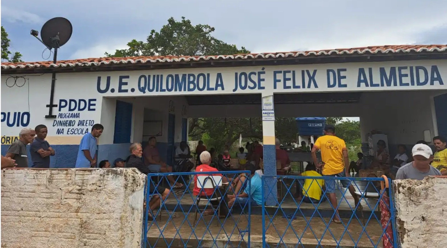 grupo de pessoas, de várias idades, espalhadas pelo hall de entrada da escola Quilombola José Félix de Almeida, cujo nome está pintado em letras azuis na fachada branca da escola.