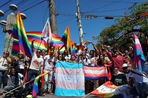Ao ar livre e sob céu azul, grupo de ativistas LGBTQIA+, posam para foto carregando ou segurando bandeiras do arco-íris e do movimento de luta das pessoas trans (com listas horizontais azul claro, rosa e branco)