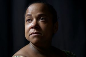 Elivaneide Nunes, mulher negra, fotografada em close, olhando para cima, em um ambiente escuro, com seus rosto parcialmente iluminado.