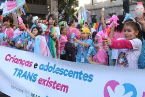 Grupo de crianças, vestidas com roupas coloridas, por trás de uma faixa com a frase crianças e adolescentes trans existem em letras azul e rosa