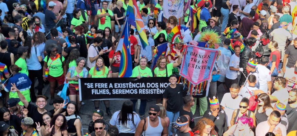 Grupo de adultos e crianças, semelhante a um bloco de carnaval, com estandarte azul, rosa e branco onde está escrito em Crianças trans existem