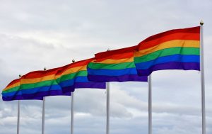 Seis bandeiras do arco-íris hasteadas, emparelhadas uma ao lado da outra, tremulando sob céu nublado.